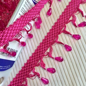 May include: Pink trim with teardrop beads. The trim is 12 yards long and made of 100% rayon. The label reads "Wrights 14465801 Teardrop 12YDS/10 100% Rayon Dust With Damp Cloth When Necessary Made in China 7065957457-0"
