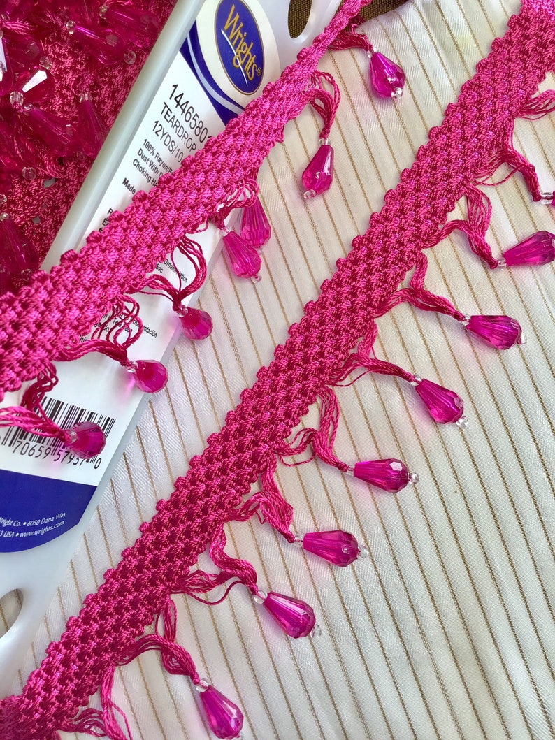 May include: Pink trim with teardrop beads. The trim is 12 yards long and made of 100% rayon. The label reads "Wrights 14465801 Teardrop 12YDS/10 100% Rayon Dust With Damp Cloth When Necessary Made in China 7065957457-0"
