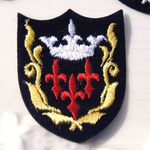 May include: A navy blue embroidered patch in the shape of a shield with a gold crown, three red fleurs-de-lis, and gold scrollwork.