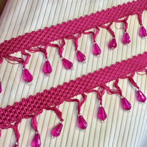 May include: Pink trim with a woven texture and dangling pink beads. The trim has a fringe detail.