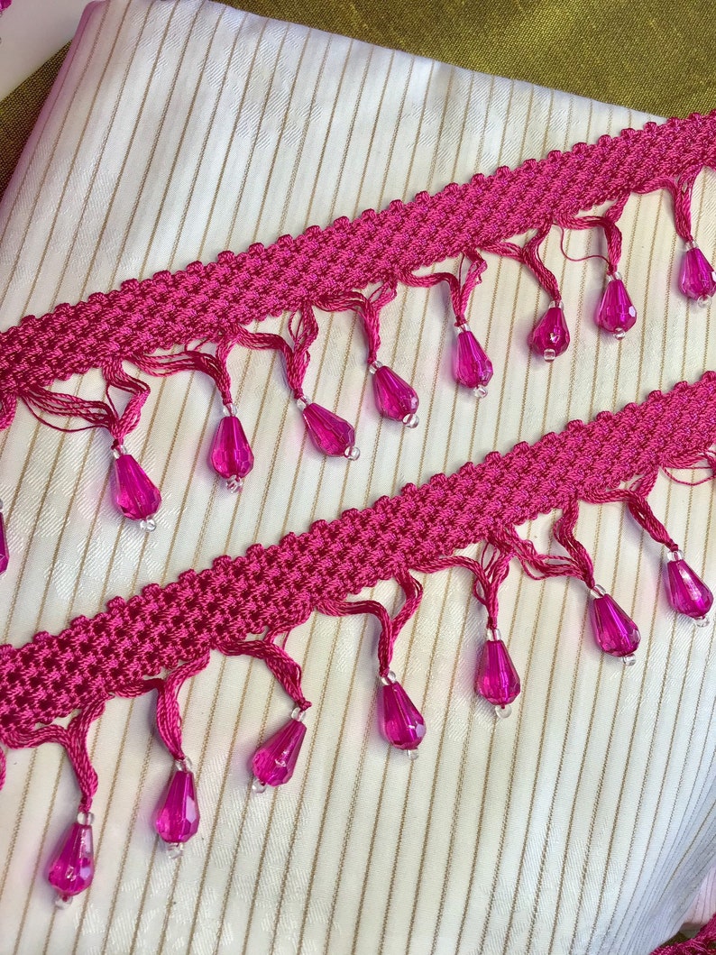 May include: Pink trim with a woven texture and dangling pink beads. The trim has a fringe detail.