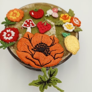 Puede incluir: Primer plano de una colección de flores, frutas y verduras de fieltro bordadas. Las flores son en tonos de rojo, amarillo y blanco. Las frutas y verduras son en tonos de rojo, naranja y amarillo. El bordado es detallado e intrincado.