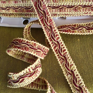 May include: A close-up of a red and cream braided trim with a woven design. The trim is made of a soft, textured fabric and has a subtle sheen.