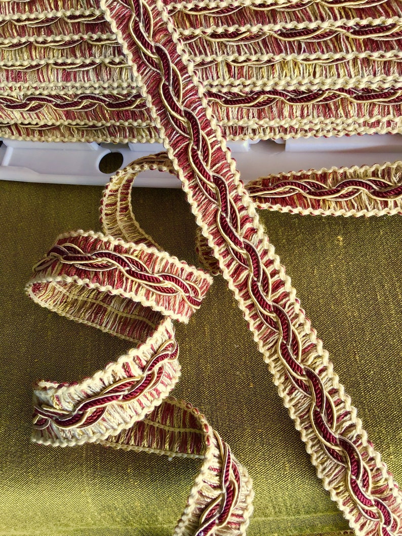 May include: A close-up of a red and cream braided trim with a woven design. The trim is made of a soft, textured fabric and has a subtle sheen.