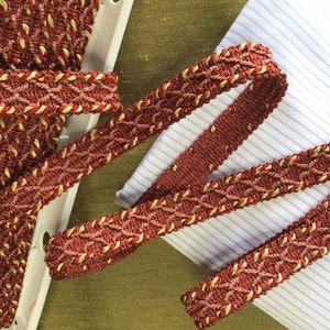 May include: Red and gold braided trim with a woven pattern. The trim is on a white and green fabric background.