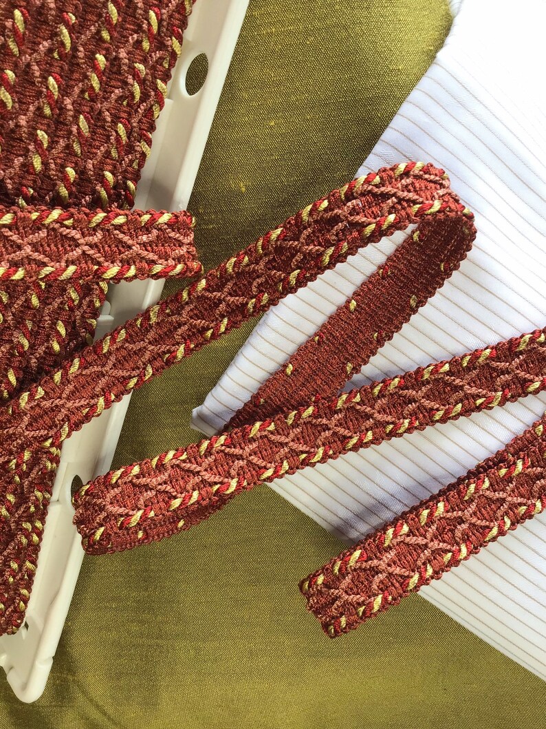 May include: Red and gold braided trim with a woven pattern. The trim is on a white and green fabric background.