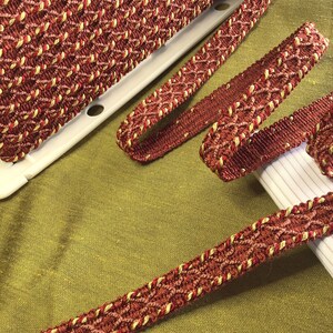 May include: Red and gold braided trim with a diamond pattern. The trim is woven with a textured, rope-like cord.