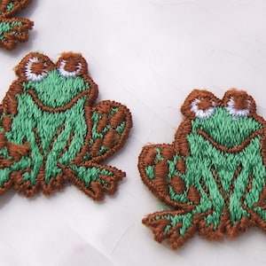 May include: Three green embroidered frog patches with brown details. The frogs have white eyes and a smiling mouth.
