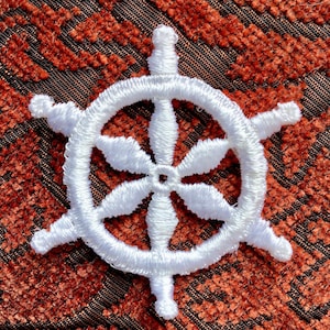 May include: White embroidered nautical wheel with eight spokes on a red textured background.