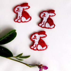 May include: Three white embroidered bunny rabbit patches with red outlines. The bunnies are sitting with their ears up and their tails behind them.