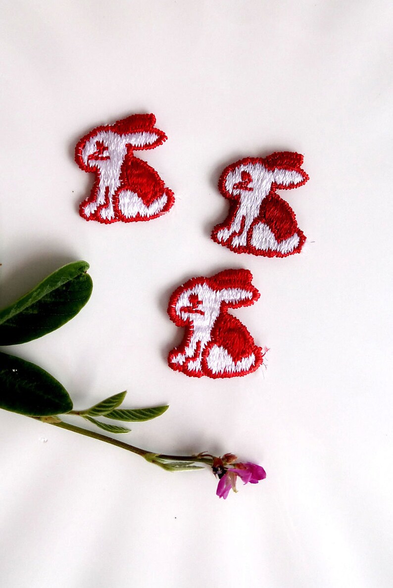May include: Three white embroidered bunny rabbit patches with red outlines. The bunnies are sitting with their ears up and their tails behind them.