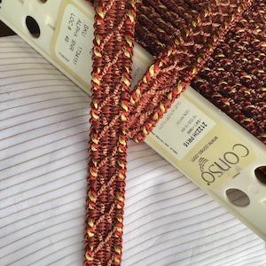 May include: A close-up of a red and gold braided trim with a diamond pattern. The trim is labeled with the SKU 1734151, ALPHA SNR, LOC # 46, and 709084.