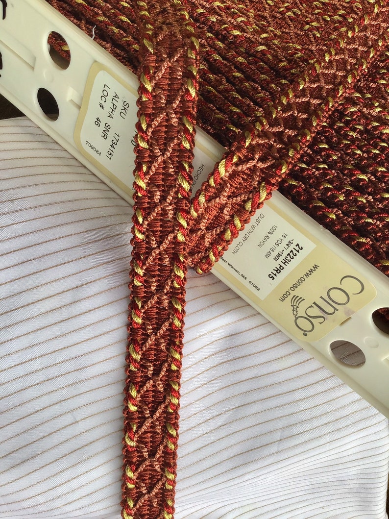 May include: A close-up of a red and gold braided trim with a diamond pattern. The trim is labeled with the SKU 1734151, ALPHA SNR, LOC # 46, and 709084.