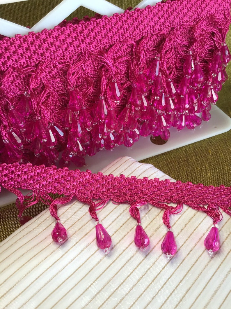May include: Pink trim with a beaded fringe. The beads are clear and faceted. The trim is shown on a white and gold striped fabric.