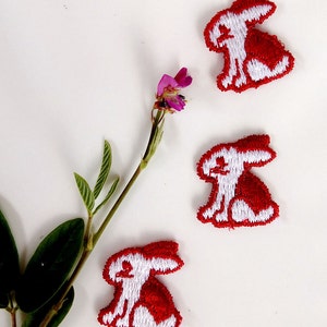 May include: Three white embroidered bunny rabbit patches on a white background. The bunnies are outlined in red and have red ears.