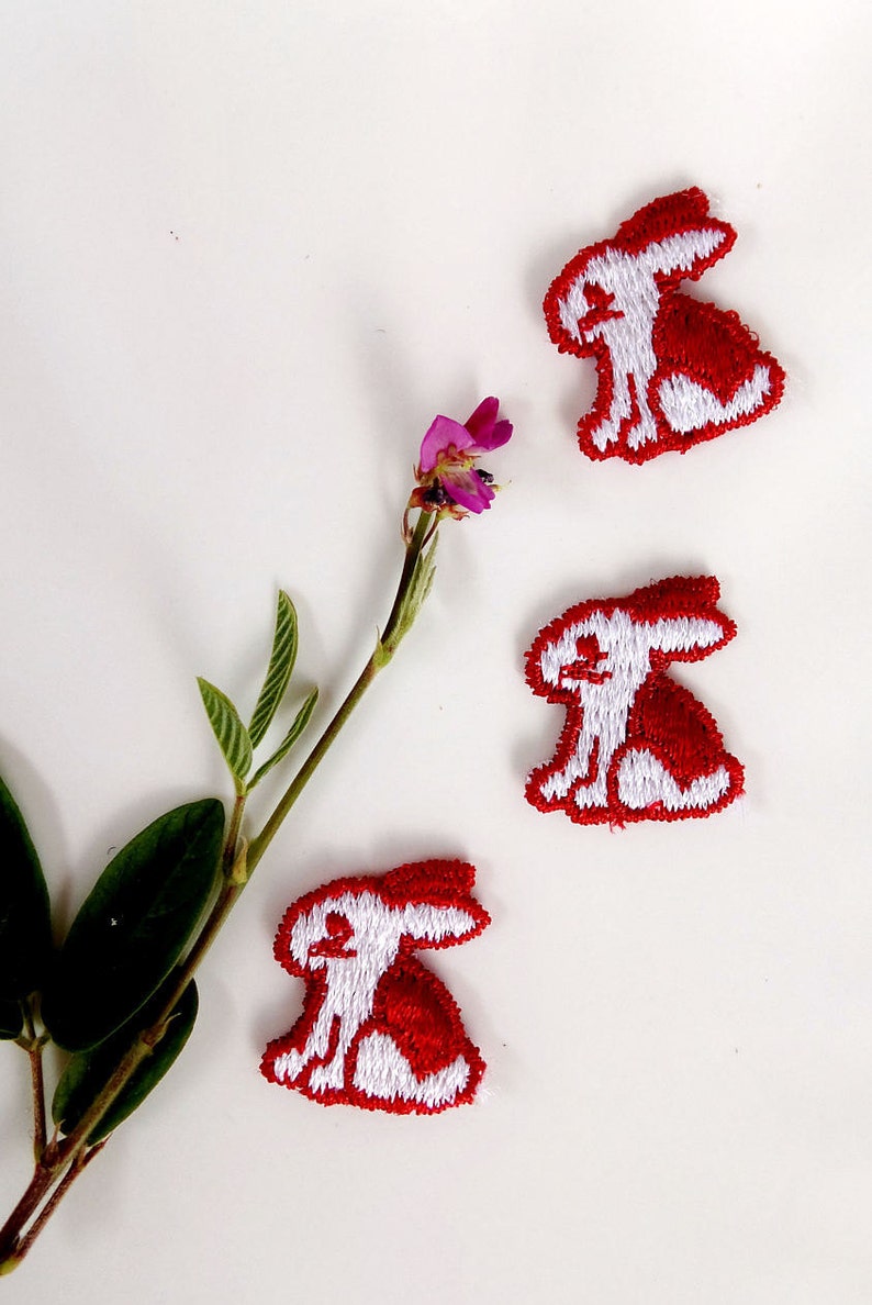 May include: Three white embroidered bunny rabbit patches on a white background. The bunnies are outlined in red and have red ears.