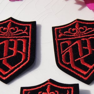 Puede incluir: Tres parches bordados rojos con respaldo de terciopelo negro. Cada parche presenta una forma de escudo con una corona y las letras "WB" en una fuente estilizada.