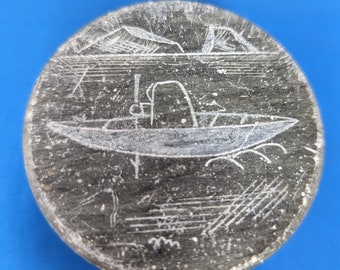 Può includere: Un'incisione rotonda in pietra grigia raffigurante una barca sull'acqua con montagne sullo sfondo. La barca ha una persona al suo interno. L'incisione è posta su uno sfondo blu.