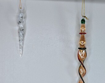 May include: Two glass Christmas decorations suspended against a white backdrop. One is clear with a spiral pattern, the other is brown and red with a twisted design. Both are hung by gold cords.