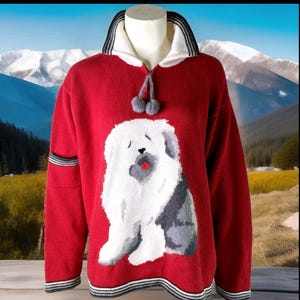 Peut inclure: Pull rouge avec un grand motif de Bobtail blanc et gris. Le pull a un col blanc avec des rayures noires, un cordon de serrage avec des pompons gris et des poignets et un ourlet rayés noir et blanc. Le fond montre un paysage de montagne.