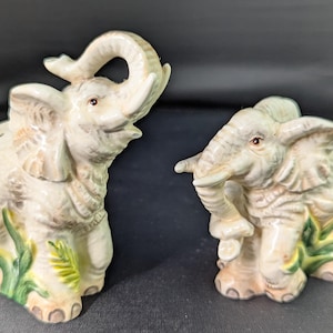 May include: Two ceramic elephant figurines. One elephant has its trunk raised, the other has its trunk lowered. Both are off-white with brown and green accents. The figurines are likely salt and pepper shakers.