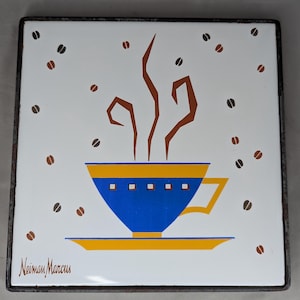 May include: Square ceramic tile with a coffee cup design. The cup is blue with yellow accents, and steam rises from it. Coffee beans are scattered around the cup on a white background. The tile has a dark metal frame and the text "Neiman Marcus" is in the bottom left corner.