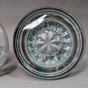 Op de afbeelding: Een heldere glazen deurknop met een decoratief, verhoogd, bloemmotief in het midden. De deurknop heeft een afgeschuinde rand en een zilverkleurige rand.