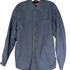 May include: A denim shirt jacket in a medium blue wash. The jacket has a button-down front, two chest pockets, and buttoned cuffs. The collar is a classic point collar. The brand name "Wrangler" is visible on the collar.