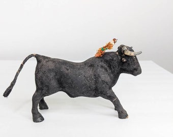 May include: A black bull figurine with a brown and green tassel on its head. The bull has a long tail and is standing on a white surface.
