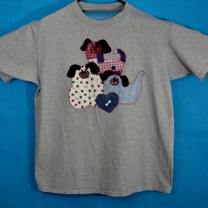May include: A gray t-shirt featuring appliqued dog designs. The dogs are made from various patterned fabrics, including red and white checkered, and paw print patterns. A denim heart with a bone is also part of the design.