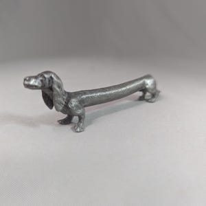Op de afbeelding: Een zilverkleurig teckelbeeldje, gedetailleerd en realistisch vormgegeven. De hond wordt staand afgebeeld, met zijn lange lichaam en korte poten duidelijk zichtbaar. Het beeldje is gemaakt van een metalen materiaal en het oppervlak heeft een textuur.
