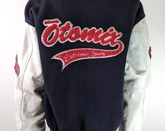 Op de afbeelding: Een donkerblauw en wit varsity jack met leren mouwen. De achterkant van het jack heeft een rood geborduurd logo met de tekst "Otomix Extreme Sports".