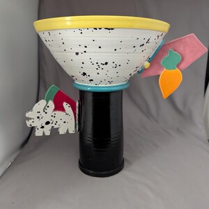 May include: A decorative ceramic bowl with a black base and a white speckled bowl. The rim is yellow, and the base has a turquoise accent. The bowl has two side decorations, one with a Dalmatian-like pattern and the other with a carrot.