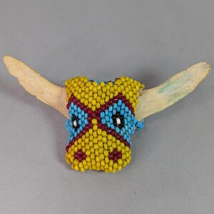 May include: A decorative object featuring a steer skull shape adorned with small glass beads in yellow, light blue, and dark red. The horns are made of a natural, off-white bone material, extending outward from the beaded center piece.