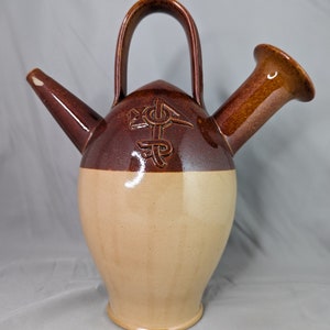 May include: A brown and beige ceramic pitcher with a handle and two spouts. The pitcher has a decorative design on the front.