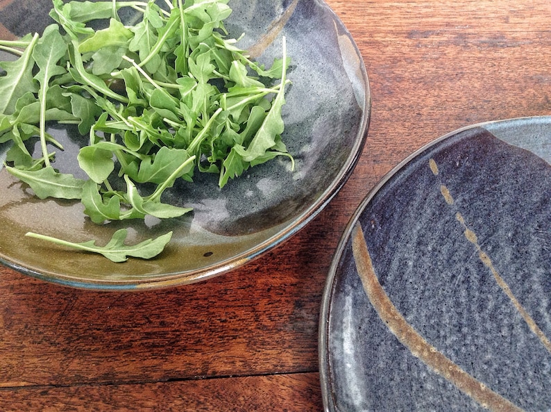 May include: Two ceramic plates, one with a dark blue glaze and gold accents, and the other with a dark green glaze and a light blue rim. The plate with the green glaze has a pile of fresh arugula on it.