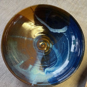 May include: A large, round ceramic bowl with a blue, brown, and white glaze. The bowl has a unique, swirling pattern.