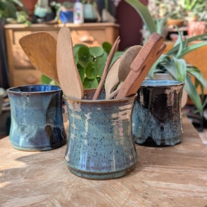 May include: Three ceramic utensil holders in shades of blue and brown sit on a wooden table. The holders contain wooden spoons and spatulas. The holders have a textured, glazed finish, and are of varying heights.