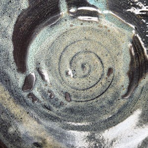 Puede incluir: Primer plano de un cuenco de cer&aacute;mica con un dise&ntilde;o en espiral. El cuenco presenta una superficie texturizada con una paleta de colores gris, negro y verde p&aacute;lido. La espiral es oscura y el cuenco tiene un acabado brillante.