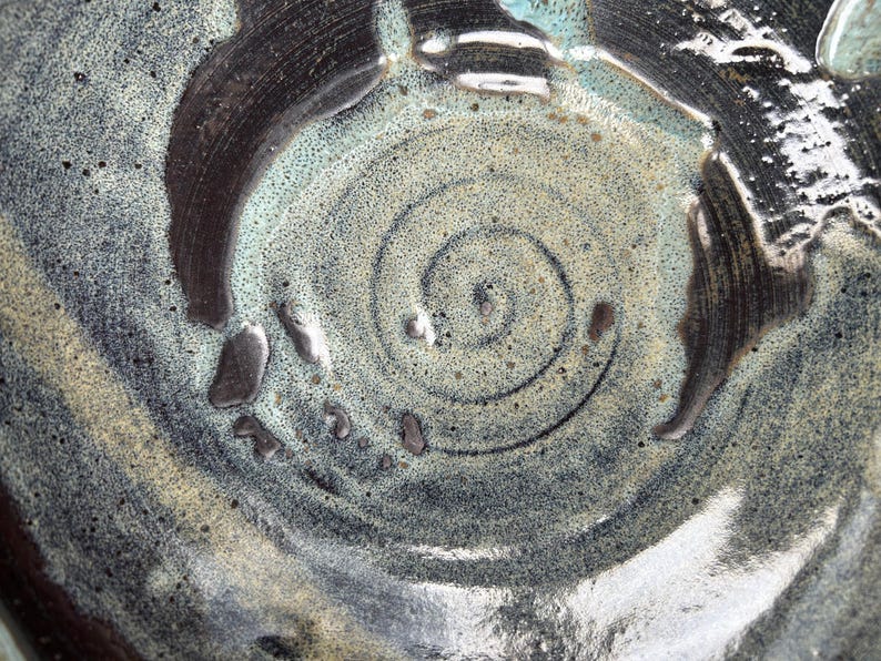 Puede incluir: Primer plano de un cuenco de cer&aacute;mica con un dise&ntilde;o en espiral. El cuenco presenta una superficie texturizada con una paleta de colores gris, negro y verde p&aacute;lido. La espiral es oscura y el cuenco tiene un acabado brillante.