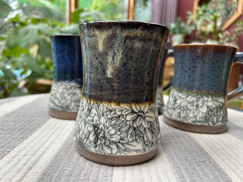 May include: Handmade ceramic mugs with a floral design. The mugs have a dark, speckled glaze on the upper portion, transitioning to a white floral pattern on the lower half. The mugs are on a striped surface.