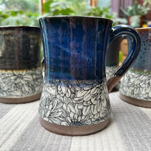 May include: A collection of handmade ceramic mugs with floral designs. The mugs feature a variety of colors, including blue, brown, and black, with white floral patterns on the lower half. Each mug has a handle and a rustic, textured base.