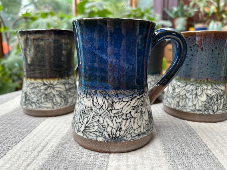 May include: A collection of handmade ceramic mugs with floral designs. The mugs feature a variety of colors, including blue, brown, and black, with white floral patterns on the lower half. Each mug has a handle and a rustic, textured base.
