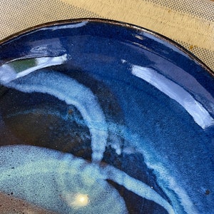 May include: A blue ceramic plate with a white glaze design. The plate has a dark blue rim and a light blue glaze that swirls and flows across the surface.