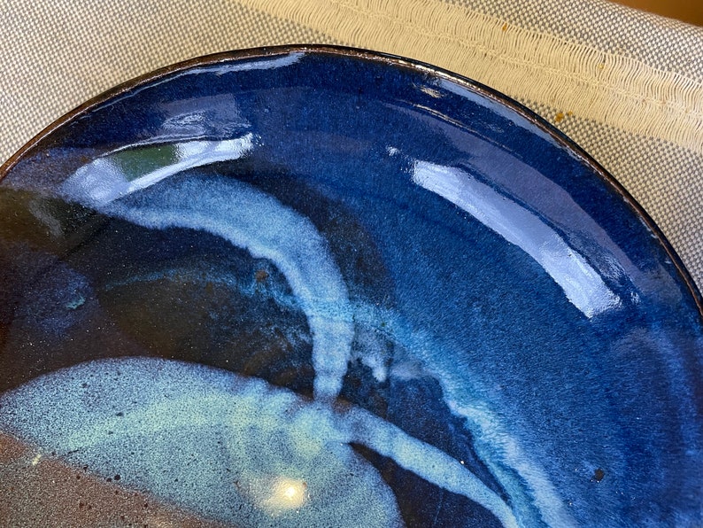 May include: A blue ceramic plate with a white glaze design. The plate has a dark blue rim and a light blue glaze that swirls and flows across the surface.