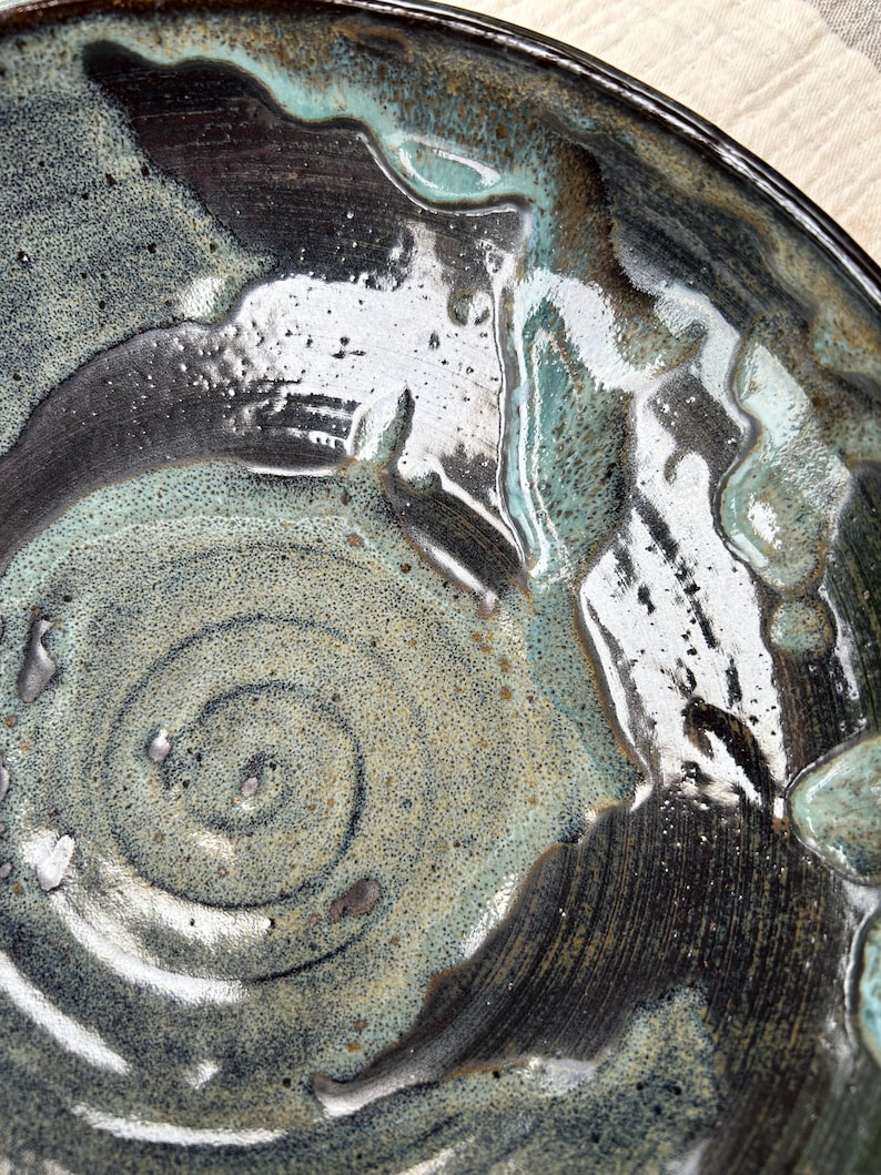 Puede incluir: Primer plano de un cuenco de cer&aacute;mica hecho a mano con un esmalte &uacute;nico. El cuenco presenta un dise&ntilde;o en espiral en el centro y est&aacute; acabado con una mezcla de colores turquesa, negro y blanco. La superficie brillante refleja la luz.