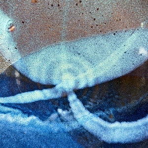 May include: Close-up of a ceramic surface with a blue and white glaze. The glaze has a textured, almost bubbly appearance. The surface is dark blue in the center, with lighter blue and white streaks radiating outwards.