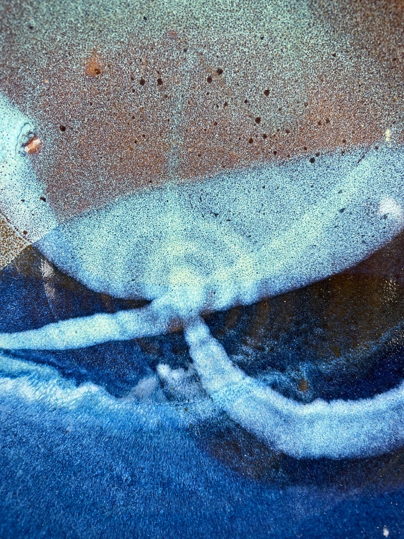 May include: Close-up of a ceramic surface with a blue and white glaze. The glaze has a textured, almost bubbly appearance. The surface is dark blue in the center, with lighter blue and white streaks radiating outwards.