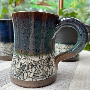 May include: Ceramic mugs with a floral design. The mugs have a dark blue and brown glaze with a white floral pattern on the lower half. The handles are curved and the mugs are sitting on a striped surface.