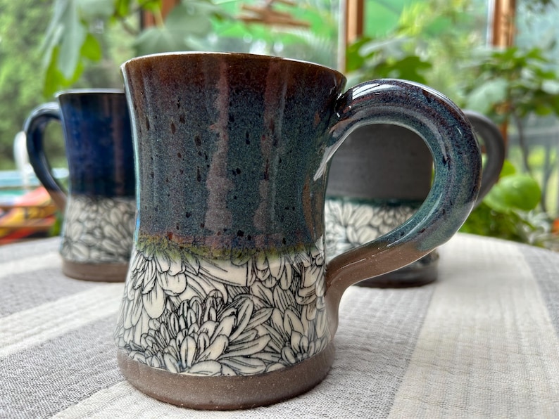 May include: Ceramic mugs with a floral design. The mugs have a dark blue and brown glaze with a white floral pattern on the lower half. The handles are curved and the mugs are sitting on a striped surface.